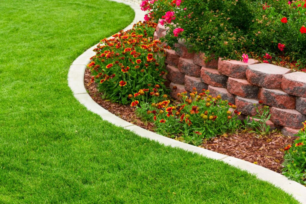Vibrant flower bed with red and yellow blooms curves along a manicured green lawn, bordered by a decorative stone wall and mulch.
